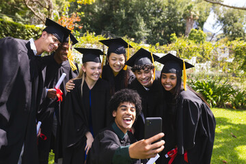 Diverse graduates in caps and gowns taking selfie, celebrating success in garden together