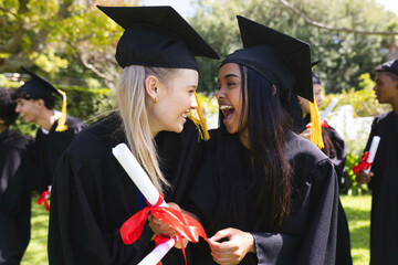 Graduating diverse students in caps and gowns celebrating with diplomas in garden, smiling