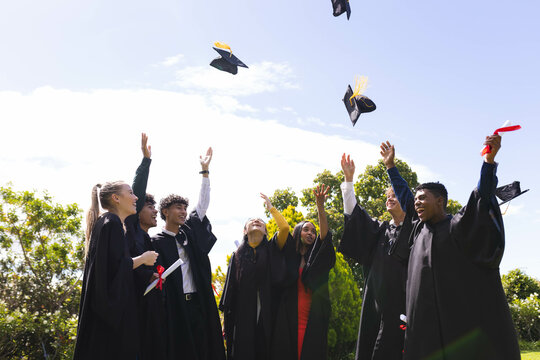 Graduating diverse students joyfully throwing caps in air, celebrating achievement in garden