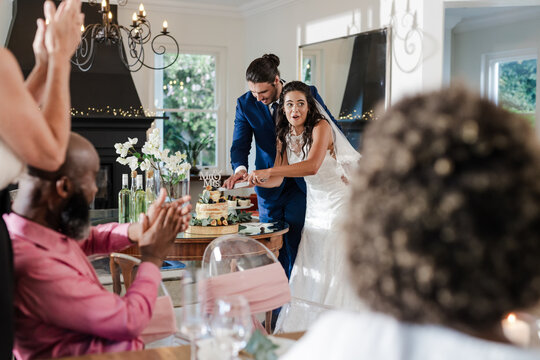 Bride and groom cutting wedding cake while guests applaud in celebration - Powered by Adobe