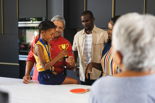 Granddaughter celebrating with diverse family, holding trophy in kitchen, feeling proud, at home