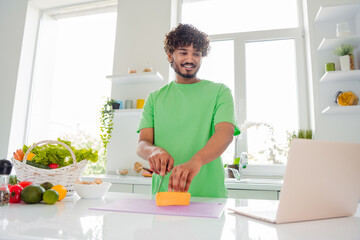 Young smiling man cooking in a bright modern kitchen with fresh vegetables, preparing a meal while referencing a recipe on his computer