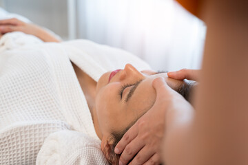 Asian serene female receiving acial massage at spa salon.  Woman in white robe getting gently massage temples on forehead for pampering and stress relief. Spa skin and body care concept