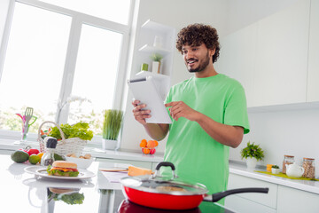 Young man in a bright kitchen using a tablet while preparing a meal, surrounded by fresh ingredients, enjoying cooking and lifestyle.