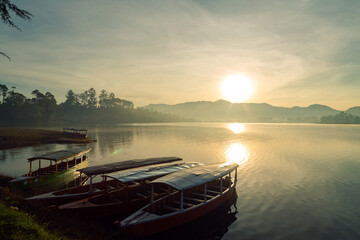 Obraz premium Sun rises over a calm misty lake with boats docked on the shore
