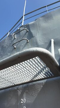 Low-angle view of a metal safety platform with grated surface and vertical ladder attached to the side of a ship, under bright daylight.