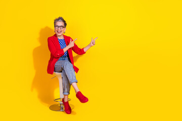Stylish female model in red jacket pointing playfully while seated against vibrant yellow background