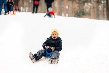 Happy child sliding down snowy hill on plastic sled in winter forest