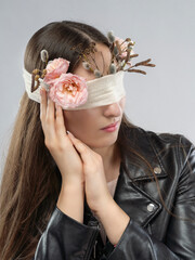 A girl with closed eyes, adorned with a blindfold decorated with flowers and branches, wearing a leather jacket. A conceptual portrait with elements of symbolism, mystery, and natural aesthetics.