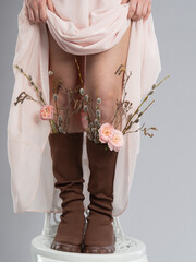 A conceptual shot of a girl standing on a white chair, wearing a flowing pastel dress and a leather jacket. Her boots are decorated with flowers and branches, creating an artistic and symbolic image