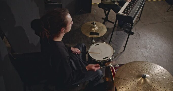 Drummer practicing in a dark studio with a minimalist drum kit. A keyboard player can be seen in the background.