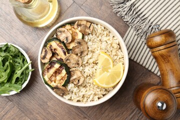 Delicious brown rice with mushrooms, zucchini and lemon slices served on wooden table, flat lay