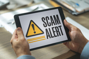 Close-up of hands holding tablet displaying scam alert warning sign, symbolizing online security risk and fraud prevention, important for digital safety awareness
