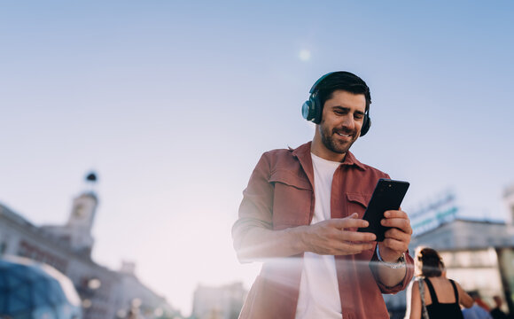 young male traveler in engages with smartphone app in bustling city square. With headphones on, he enjoys digital content while exploring European destination, combining leisure and technology on go