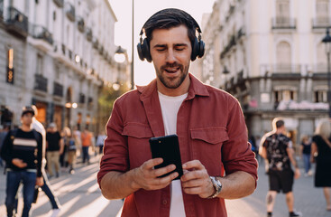 stylish man wearing wireless headphones walks through a lively city street at sunset, engaging with...