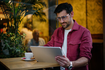 Male student studies online in relaxed café environment, engaging with his tablet while drinking coffee. Man combines education and lifestyle, utilizing digital tools for efficient learning and growth