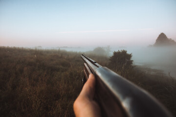 Hunter hand holding shotgun aiming during foggy morning on rural field during hunting season as...