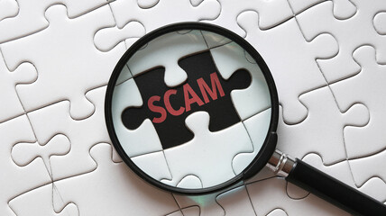 Magnifying glass focuses on a missing jigsaw puzzle piece with "scam" written on it, amidst a field of white puzzle pieces, symbolizing the detection and investigation of fraudulent activities