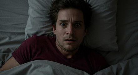 Obraz premium A terrified man jolted awake in the dead of night, his eyes wide with fear, under muted bedroom lighting.