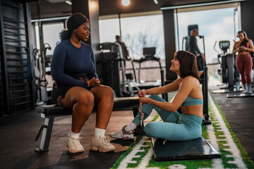 Two diverse female athletes are taking a break from their training, sitting and chatting on a weight bench and a mat in a modern gym, fostering camaraderie and support within their fitness journey