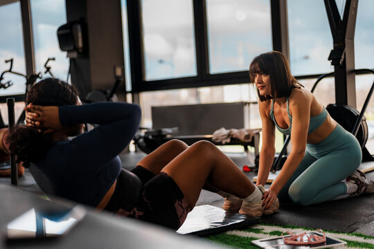 Determined overweight woman performing sit ups with assistance from personal trainer, focusing on core strength and fitness goals in modern gym setting