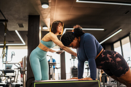Overweight woman doing push ups with personal trainer assisting and controlling her posture in a gym, focusing on healthy lifestyle and overcoming challenges