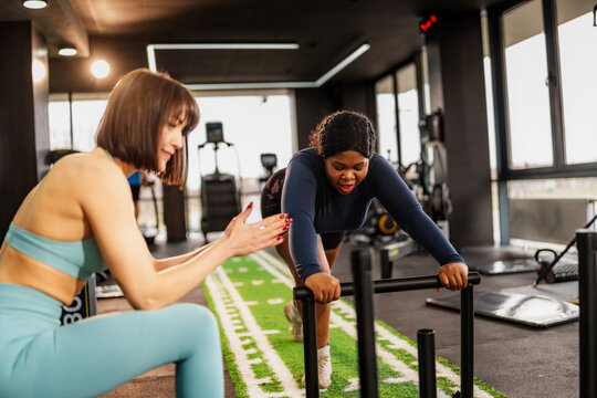 Overweight woman is pushing a weight sled on artificial turf in a gym, following the instructions of her personal trainer, who is clapping and encouraging her