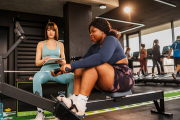 Overweight woman working hard on a rowing machine with a personal trainer documenting progress in a bustling gym, highlighting a dedicated fitness journey focused on health and wellness