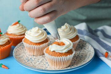 Woman squeezing whipped cream onto delicious carrot cupcakes at blue table, closeup