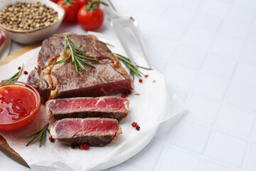 Pieces of delicious beef meat, spices, sauce and fork on white tiled table, closeup. Space for text
