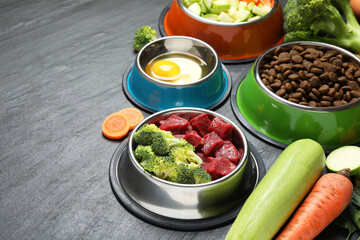 Natural and dry pet food in feeding bowls among products on dark textured table, closeup. Space for text
