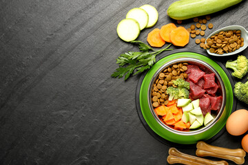 Natural and dry pet food in feeding bowl among products on dark textured table, flat lay. Space for text