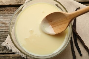 Taking tasty vanilla condensed milk with spoon from bowl on wooden table, flat lay