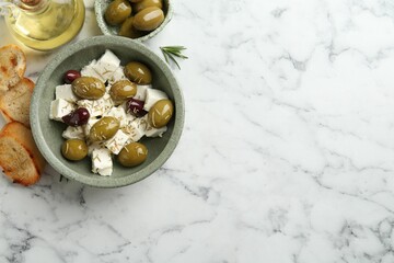 Marinated olives with feta cheese, bread pieces, oil and rosemary on white marble table, flat lay. Space for text