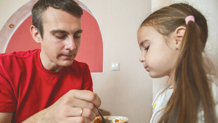 Cheerful small girl daughter and her handsome young dad spending time together at home. Father feeding his little daughter.