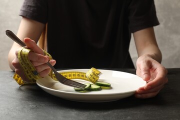 Bulimia. Woman with measuring tape eating pieces of cucumber at dark textured table, closeup