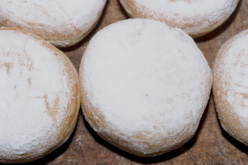 Okrągłe pączki posypane cukrem pudrem leżące na drewnianym blacie w Tłusty Czwartek.  Pączki są równomiernie rozmieszczone, co podkreśla ich jednolity kształt i miękki wygląd. Drewno ma fakturę 