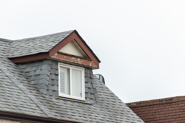 Detailed view of a residential roof dormer featuring grey shingles, a white-framed window, and peeling paint on the trim.
