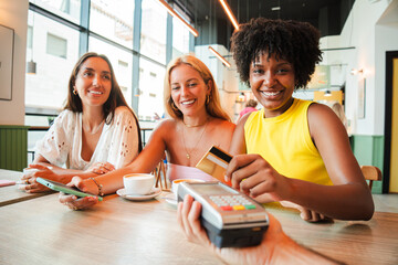 Three cheerful women sharing a joyful moment while completing a payment transaction at a trendy cafe with a contactless credit card, celebrating friendship and connection over their favorite beverages