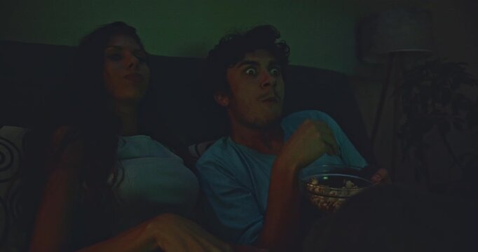 Couple enjoys a scary movie night at home on the couch with popcorn. She flinches and laughs as the light from the TV flashes across their faces.