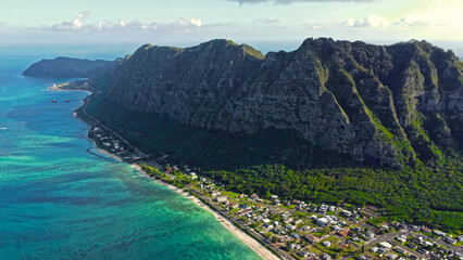 Coastal Town Beneath Rugged Hawaiian Cliffs. Aerial view of a tropical seaside town nestled at the...