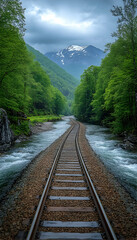 Fototapeta premium Scenic railway tracks between a river & forest with a mountain backdrop under a cloudy sky