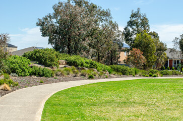 A curved concrete pathway winds through a well-maintained suburban park in Tarneit, Melbourne, bordered by a lush green lawn and native landscaped garden beds with shrubs and trees.