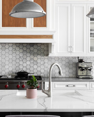A kitchen detail with metal lights hanging above a large marble island, stainless faucet on a white farmhouse sink, and a marble hexagon tiled backsplash with white cabinets.