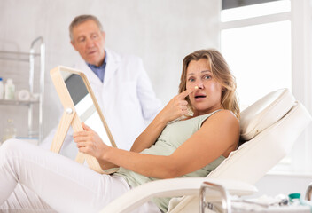 Obraz premium Upset adult woman examines her face in mirror after cosmetic procedure