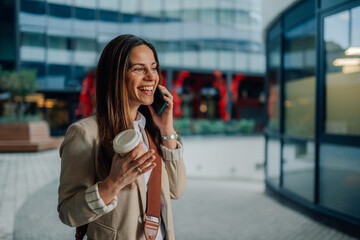 Fototapeta premium Businesswoman talking on phone and holding coffee outside office
