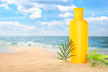 Bottle of sunscreen product and green tropical leaves on sandy beach near ocean