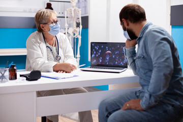 Senior doctor seated at desk with laptop displaying virus cells and discussing with male patient in modern clinic. Old female physician and young man using digital device during medical consultation.