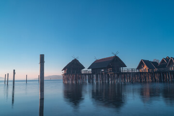 Tranquil morning at a lakeside pier with wooden houses reflecting on calm waters in a serene landscape