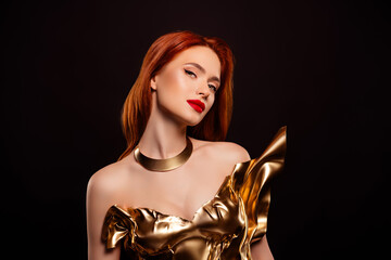 Passionate woman with red hair in golden dress posing against dark background with a glamorous and...
